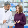 Front view of female customer consulting with chemist, holding blister pack of pills. Bearded pharmacist wearing lab coat, helping young woman in pharmacy with medicaments, explaining instruction.