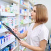 Female pharmacist standing near shelves with medicaments in drugstore. Side view of woman in white lab coat holding folder in hands. Specialist working in pharmaceutical store. Medicine concept.