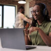 Smiling woman with wireless headphones watching entertainment show on laptop while sitting at desk. African american freelancer looking at funny social media content on portable computer.